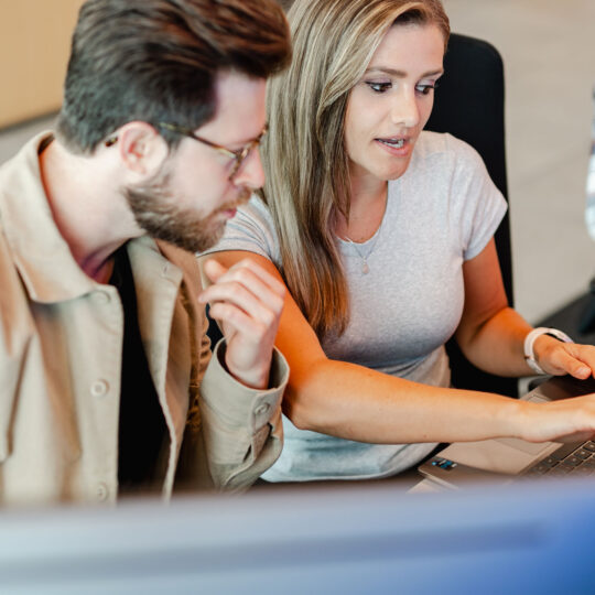 Two coworkers sitting at a desk together, reviewing a file on a laptop