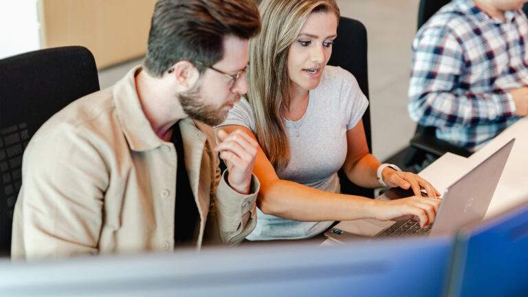 Two coworkers sitting at a desk together, reviewing a file on a laptop